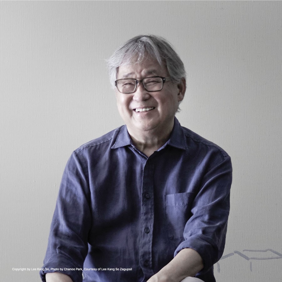전시 공식 작가 사진 — 밝은 회색 벽 앞에서 남색 셔츠를 입은 이강소가 안경을 쓰고 웃고 있습니다.